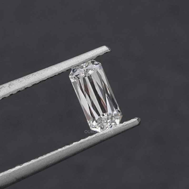 May include: A clear, rectangular emerald cut diamond gemstone held in tweezers. The diamond has a brilliant, faceted surface.