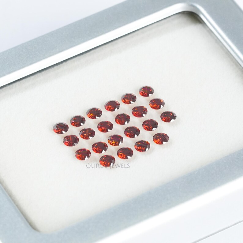 Puede incluir: Una disposici&oacute;n en forma de coraz&oacute;n de 25 peque&ntilde;as piedras preciosas redondas rojas en una caja blanca. Las piedras preciosas est&aacute;n dispuestas en forma de coraz&oacute;n con la punta del coraz&oacute;n hacia arriba. La caja es blanca con un borde plateado.