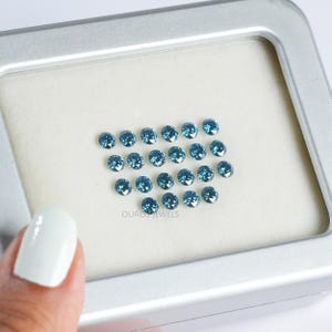 May include: A close-up view of a collection of loose blue diamonds in a white box. The diamonds are arranged in a heart shape. The box is labelled "OUROS JEWELS".