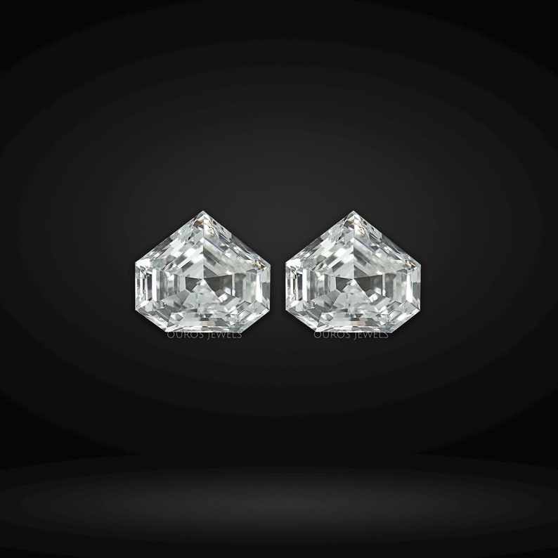 May include: Two loose, colorless, pentagon-shaped diamonds with a brilliant cut, featuring multiple facets that reflect light. Each stone displays a sharp, pointed top and a flat base, with the small text OUROS JEWELS printed at the bottom.