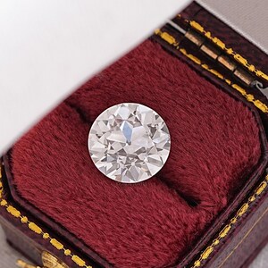 May include: A large, round, clear diamond gemstone is displayed in a red velvet ring box.