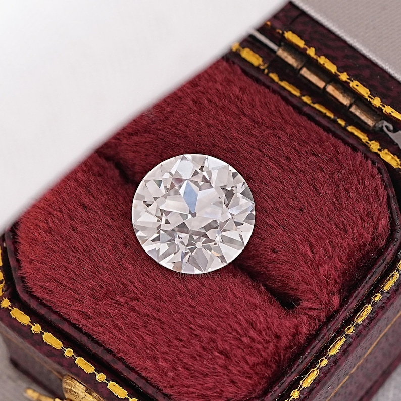 May include: A large, round, clear diamond gemstone is displayed in a red velvet ring box.