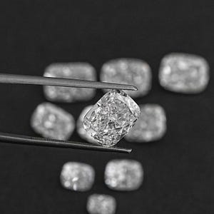 K&ouml;nnte beinhalten: Nahaufnahme eines klaren, facettierten, kissenf&ouml;rmigen Diamanten, der mit einer Pinzette gehalten wird. Der Diamant ist von anderen &auml;hnlichen Diamanten auf schwarzem Hintergrund umgeben.
