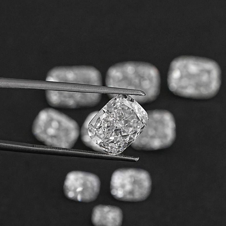 K&ouml;nnte beinhalten: Nahaufnahme eines klaren, facettierten, kissenf&ouml;rmigen Diamanten, der mit einer Pinzette gehalten wird. Der Diamant ist von anderen &auml;hnlichen Diamanten auf schwarzem Hintergrund umgeben.
