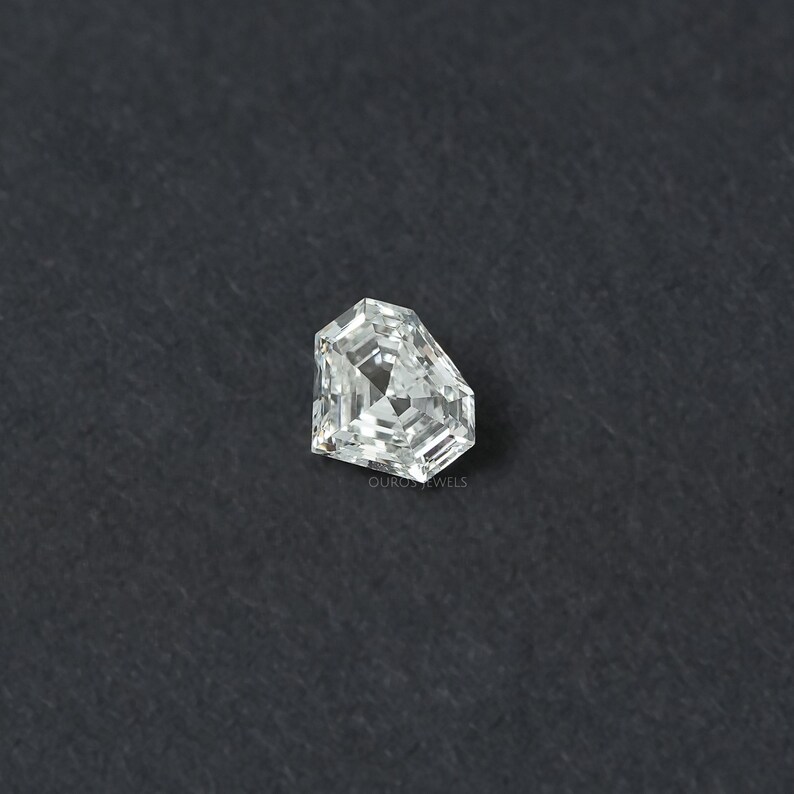 May include: A loose, colorless, pentagonal-shaped diamond with a brilliant-cut facet pattern. The gemstone features sharp, angular edges and a clear, reflective surface. The text OUROS JEWELS is printed in small, faint letters near the bottom edge.