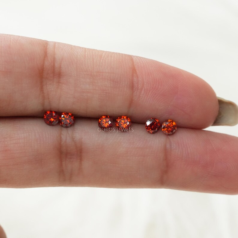 Puede incluir: Cinco piedras preciosas redondas de color rojo anaranjado, probablemente diamantes, se muestran en un dedo. Las piedras preciosas est&aacute;n dispuestas en una fila, con el texto "OUROS JEWELS" visible debajo.