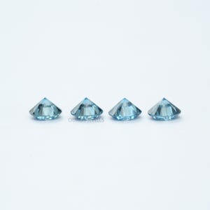 May include: Three round, faceted, light blue gemstones. The gemstones are arranged in a row on a white surface.