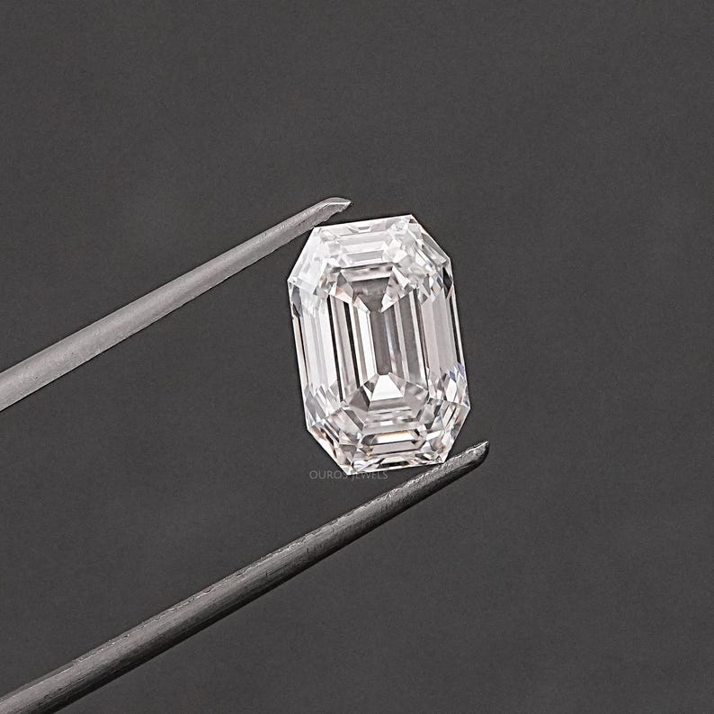 May include: A clear, rectangular emerald cut diamond gemstone held by tweezers against a grey background.