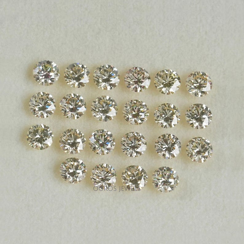 May include: A collection of 20 round, clear, faceted, loose diamonds. The diamonds are arranged in rows on a white surface.