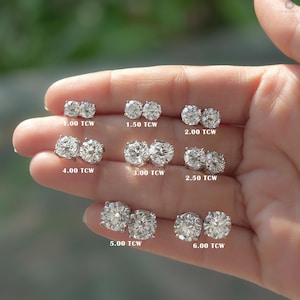 Pode incluir: Uma coleção de brincos de diamante em vários tamanhos, exibidos em uma mão. Cada par é rotulado com seu peso total em quilates (TCW), variando de 1,00 TCW a 6,00 TCW. Os brincos são de corte redondo e são cravejados em metal prateado ou branco.