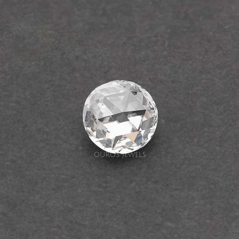 K&ouml;nnte beinhalten: Ein runder, facettierter, farbloser Edelstein im Rosenschliff mit flacher Basis und gew&ouml;lbter, mehrfach facettierter Oberseite, die Licht reflektiert. Der Text OUROS JEWELS ist in kleinen, wei&szlig;en Gro&szlig;buchstaben unter dem Stein gedruckt.