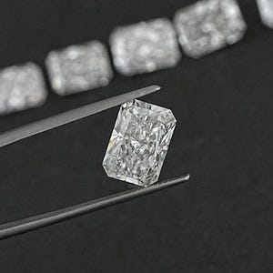 Puede incluir: Una gema de diamante transparente, rectangular y de talla esmeralda, sostenida por unas pinzas sobre un fondo negro.