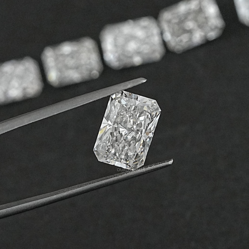 Puede incluir: Una gema de diamante transparente, rectangular y de talla esmeralda, sostenida por unas pinzas sobre un fondo negro.