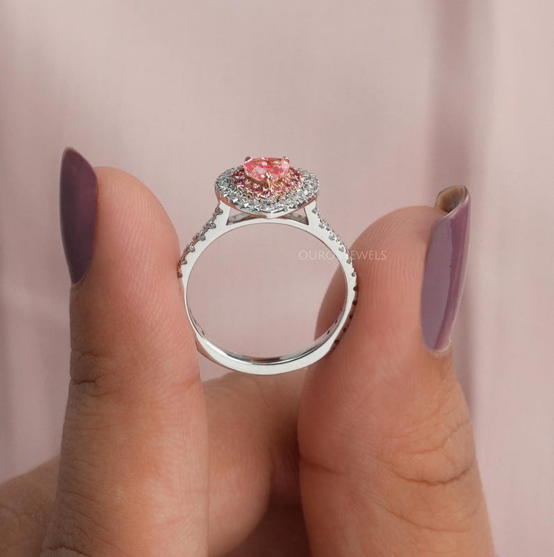 May include: A silver ring featuring a central heart-shaped pink gemstone surrounded by a halo of small white stones. The band is also set with small white stones along the sides. The text Ouro Jewels is visible on the side of the ring setting.