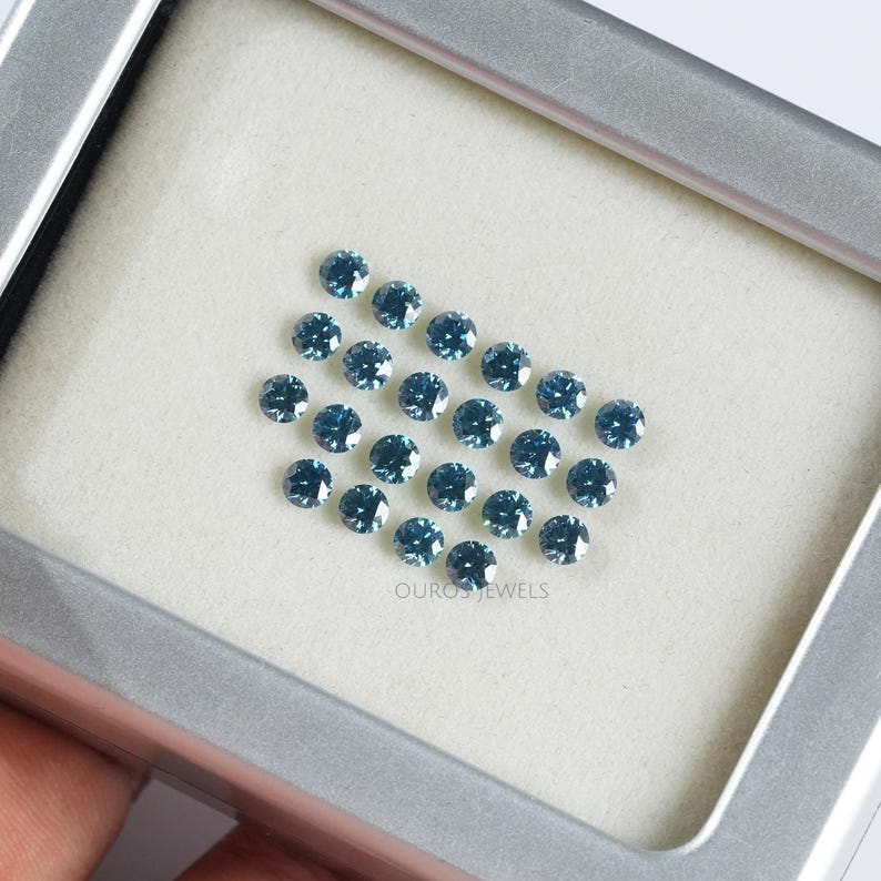 May include: A collection of 25 round, blue gemstones displayed in a white, square, framed box. The box is made of a light grey material and has a silver border. The text "OUROS JEWELS" is printed in black at the bottom of the box.
