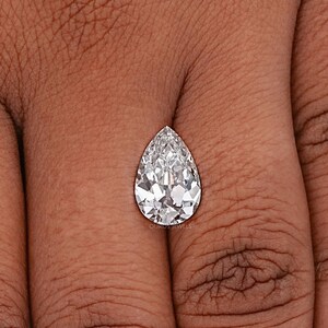 May include: A pear-shaped diamond gemstone with a clear, brilliant cut. The diamond is set on a finger, showcasing its size and sparkle.