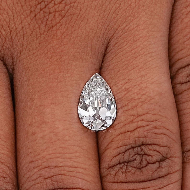 May include: A pear-shaped diamond gemstone with a clear, brilliant cut. The diamond is set on a finger, showcasing its size and sparkle.
