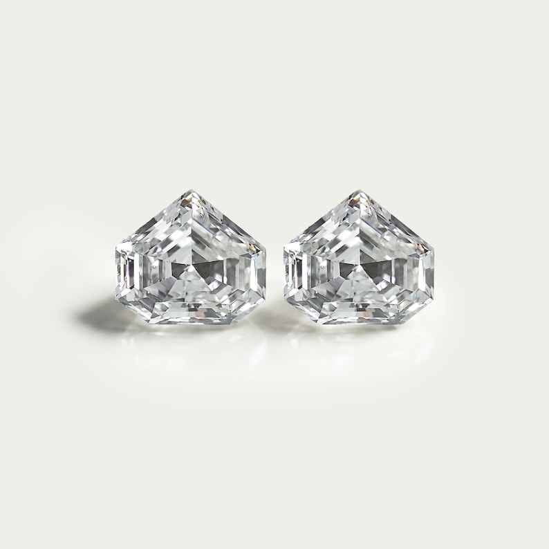 May include: Two loose, clear, colorless gemstones with a pentagonal shape and a brilliant cut. Each stone features multiple triangular and trapezoidal facets that reflect light, creating a sharp, geometric appearance with a pointed top edge.