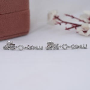 May include: A pair of silver drop earrings spelling out the word "MORE". Each letter is encrusted with small, sparkling stones. The earrings have a delicate, cursive font design. The earrings are displayed on a white surface, with a pink jewellery box in the background.