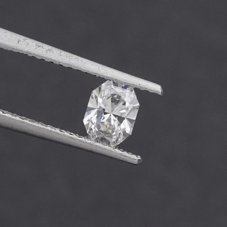 May include: A faceted, oval-cut diamond held by silver tweezers against a dark gray background. The diamond sparkles with internal reflections, showcasing its clarity and brilliance. The gem is a central focus, highlighting its cut and shape.