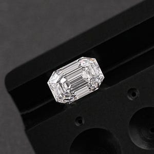 May include: A clear, rectangular emerald cut diamond gemstone is displayed in a black velvet tray. The diamond is set in a black metal setting.