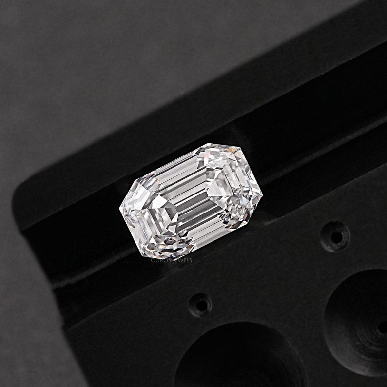 May include: A clear, rectangular emerald cut diamond gemstone is displayed in a black velvet tray. The diamond is set in a black metal setting.