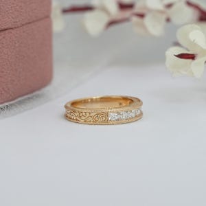 Puede incluir: Un anillo de banda dorado con un dise&ntilde;o de filigrana detallado y una fila de piedras preciosas claras de corte cuadrado. El anillo est&aacute; sobre un fondo blanco, con una caja de joyer&iacute;a rosa y flores blancas en el fondo.