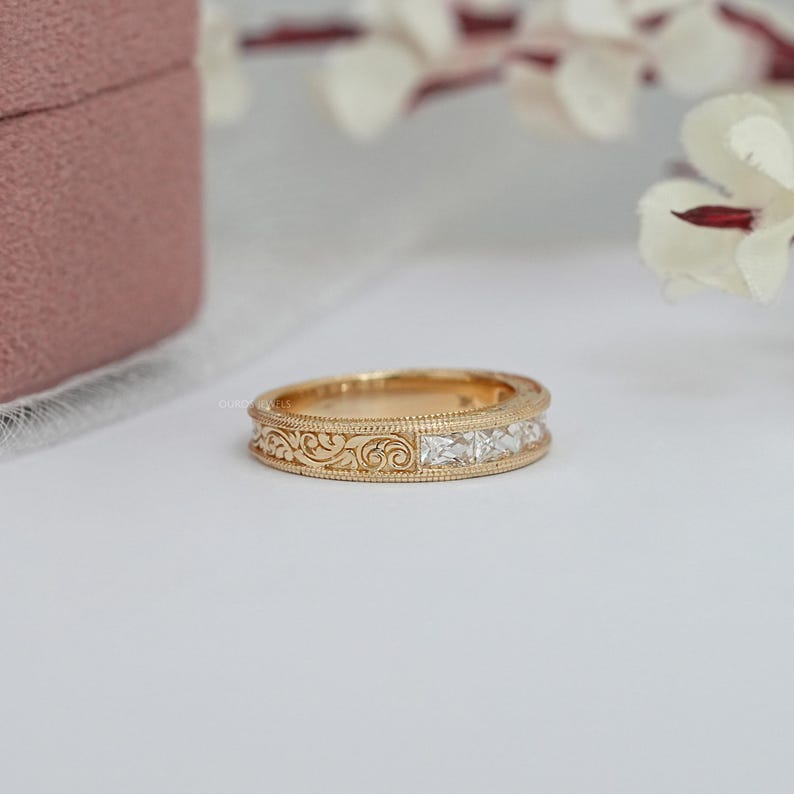 Puede incluir: Un anillo de banda dorado con un dise&ntilde;o de filigrana detallado y una fila de piedras preciosas claras de corte cuadrado. El anillo est&aacute; sobre un fondo blanco, con una caja de joyer&iacute;a rosa y flores blancas en el fondo.