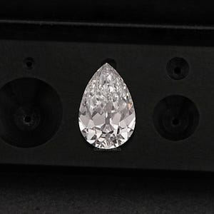 May include: A pear-shaped diamond gemstone in a black velvet box. The diamond is clear and has a brilliant cut.