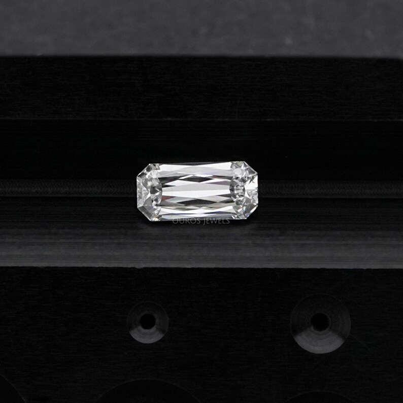 May include: A clear, rectangular emerald cut diamond gemstone on a black background. The diamond has a brilliant, faceted cut.
