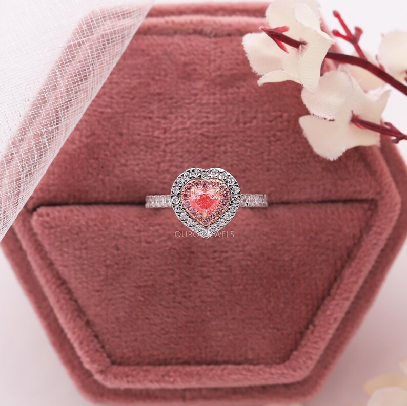 May include: A silver-toned ring featuring a central heart-shaped pink gemstone surrounded by a halo of smaller pink stones and an outer border of clear white stones. The band is thin and metallic, displaying the text OUROJEWELS on the setting.