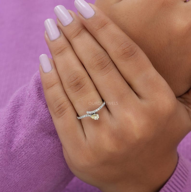 Peut inclure: Une bague bypass orn&eacute;e d'une pierre jaune en forme de poire et d'un anneau incrust&eacute; de petites pierres blanches. L'anneau en m&eacute;tal pr&eacute;sente une finition polie et se courbe autour du doigt, mettant en valeur la pierre jaune centrale et les &eacute;clats.
