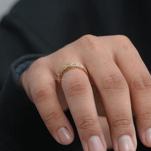 Puede incluir: Un anillo dorado con detalles intrincados y peque&ntilde;os acentos brillantes. El anillo se lleva en un dedo, mostrando su dise&ntilde;o ornamentado. La superficie del anillo tiene una apariencia texturizada, lo que aumenta su atractivo visual. El anillo probablemente est&aacute; hecho de un metal precioso.