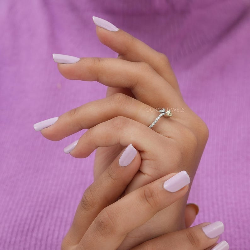 Peut inclure: Une bague en argent avec une petite pierre centrale claire et un anneau serti de minuscules pierres transparentes. La bague est port&eacute;e au doigt et le texte ORO JEWELS est visible pr&egrave;s du chaton. Le design pr&eacute;sente une structure m&eacute;tallique fine.