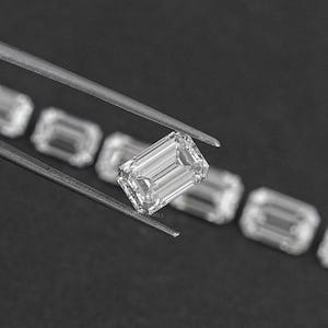 May include: A close-up of a clear, rectangular emerald-cut diamond held by tweezers. The diamond is surrounded by other smaller diamonds.