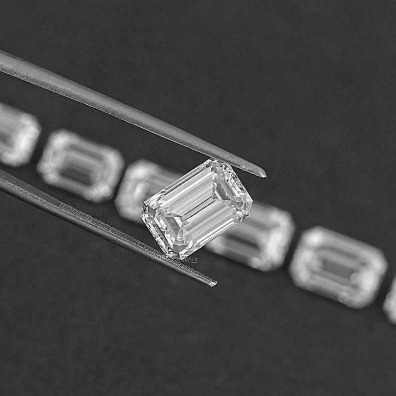 May include: A close-up of a clear, rectangular emerald-cut diamond held by tweezers. The diamond is surrounded by other smaller diamonds.