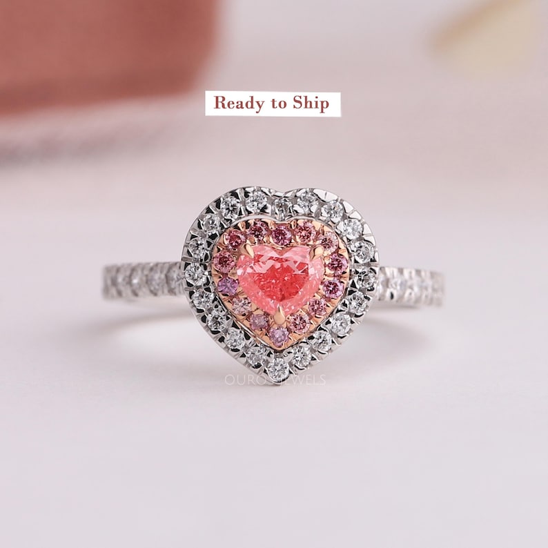 May include: A heart-shaped ring featuring a central pink heart-cut stone surrounded by a halo of smaller pink stones, an outer halo of clear white stones, and a band set with clear white stones. The text Ready to Ship is visible above the ring.