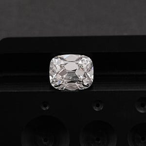 May include: A clear, cushion-cut gemstone with multiple facets, reflecting light. The gem is set against a dark, textured surface. The image is well-lit, highlighting the brilliance and clarity of the stone. The text "OUROS JEWELS" is visible.