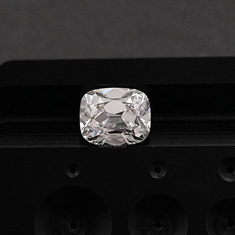 May include: A clear, cushion-cut gemstone with multiple facets, reflecting light. The gem is set against a dark, textured surface. The image is well-lit, highlighting the brilliance and clarity of the stone. The text "OUROS JEWELS" is visible.