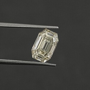 May include: A light yellow emerald cut diamond gemstone held by tweezers against a dark grey background.