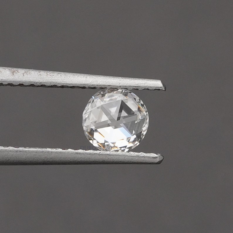 K&ouml;nnte beinhalten: Ein runder, klarer, facettierter Edelstein, der von einer silberfarbenen Metallpinzette gehalten wird. Der Stein hat einen Rosenschliff mit flacher Unterseite und einer gew&ouml;lbten, facettierten Oberseite, die Licht reflektiert.