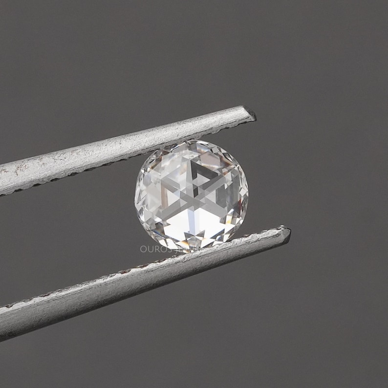 K&ouml;nnte beinhalten: Ein runder, klarer, facettierter Edelstein, der von einer Metallpinzette gehalten wird. Der Stein hat einen Rosenschliff mit einer zentralen hexagonalen Facette und strahlenf&ouml;rmigen Dreiecksfacetten. Der Text OUROS JEWELS ist sichtbar.