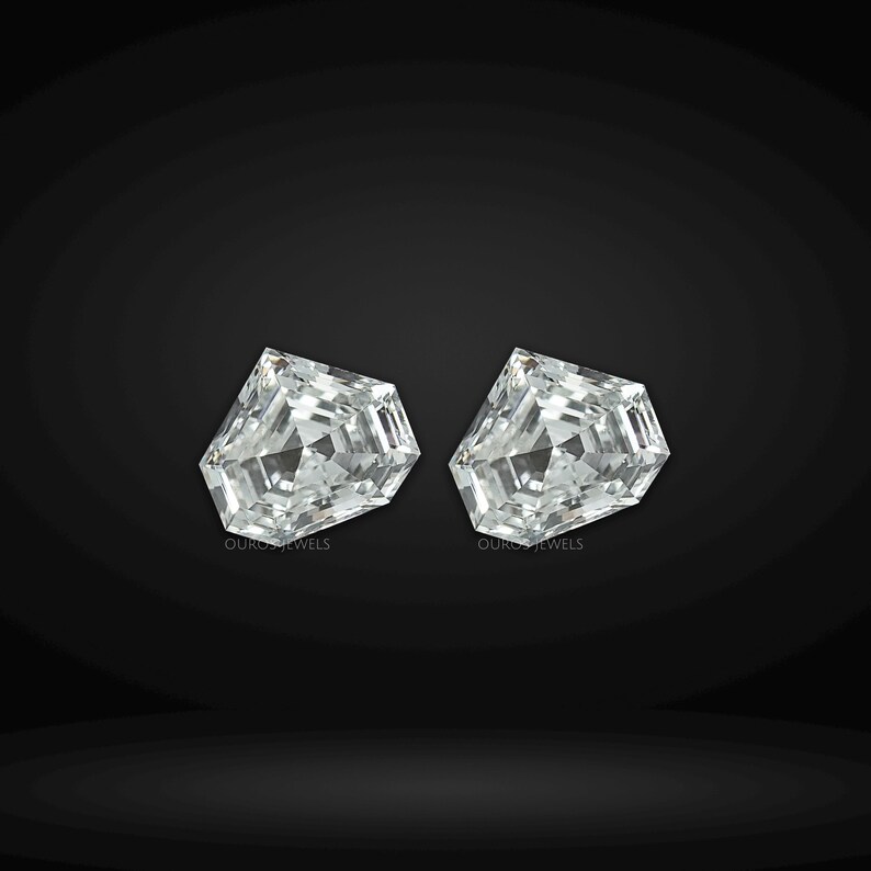 May include: Two loose, colorless, shield-cut gemstones with a faceted surface that reflects light. Each stone features a geometric, multi-sided shape with a pointed top and a flat base. The text OUROS JEWELS is printed in small letters below each stone.