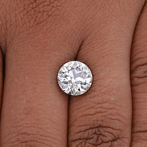 May include: A round, clear, white diamond gemstone on a finger. The diamond has many facets and is set in a simple, round setting.