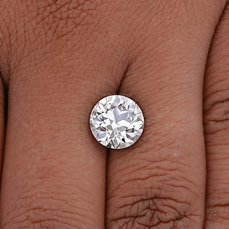 May include: A round, clear, white diamond gemstone on a finger. The diamond has many facets and is set in a simple, round setting.