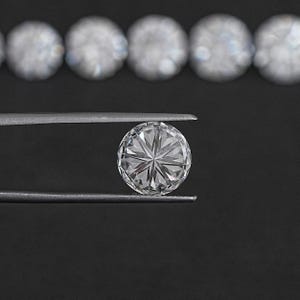 May include: A clear round diamond gemstone held by tweezers against a black background. The diamond has a star-shaped pattern.