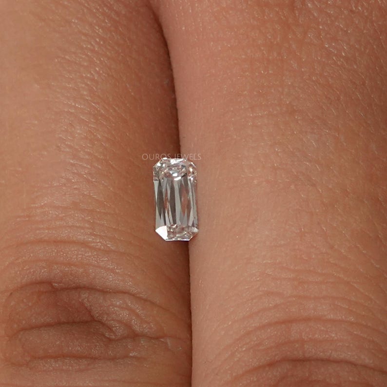 May include: A clear emerald cut diamond gemstone on a finger.
