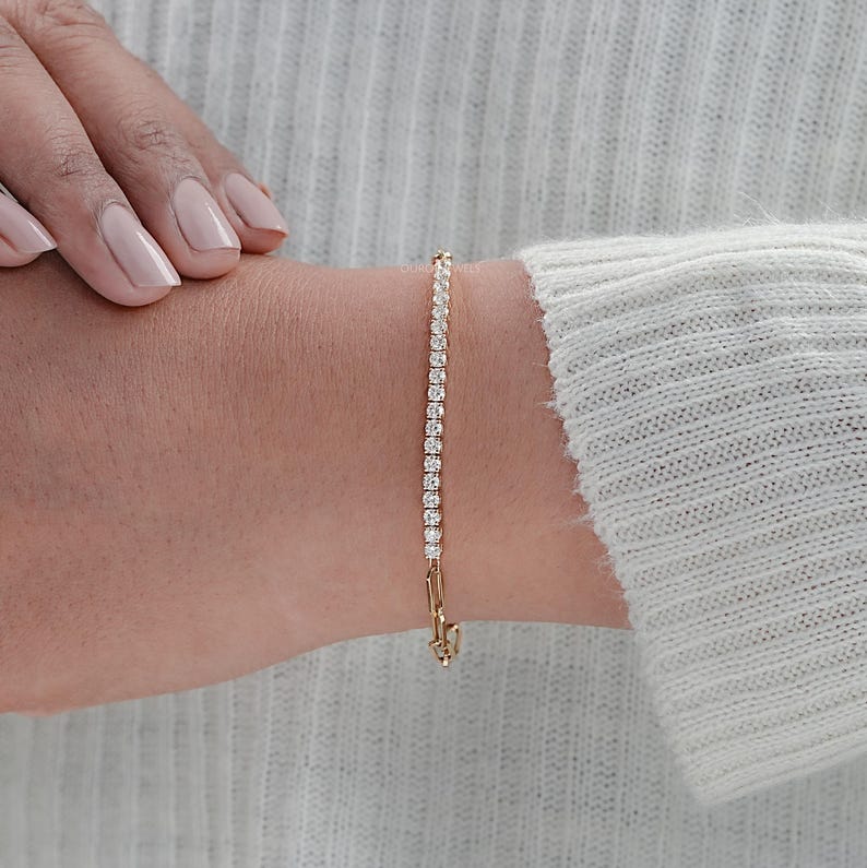 May include: A gold bracelet with a row of small, clear gemstones. The bracelet is clasped and worn on a wrist, with a light-colored sweater in the background. The gemstones are evenly spaced and reflect light.