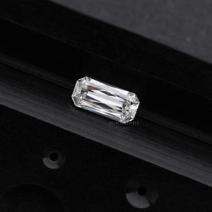 May include: A clear, rectangular emerald cut diamond gemstone on a black background. Ouros Jewels is printed below the diamond.