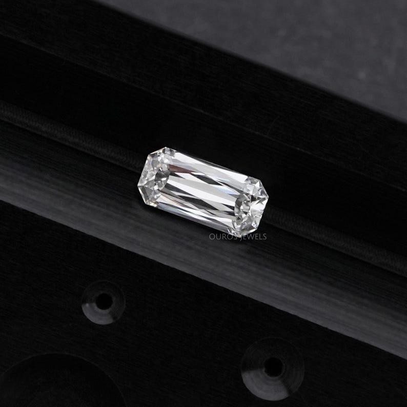 May include: A clear, rectangular emerald cut diamond gemstone on a black background. Ouros Jewels is printed below the diamond.
